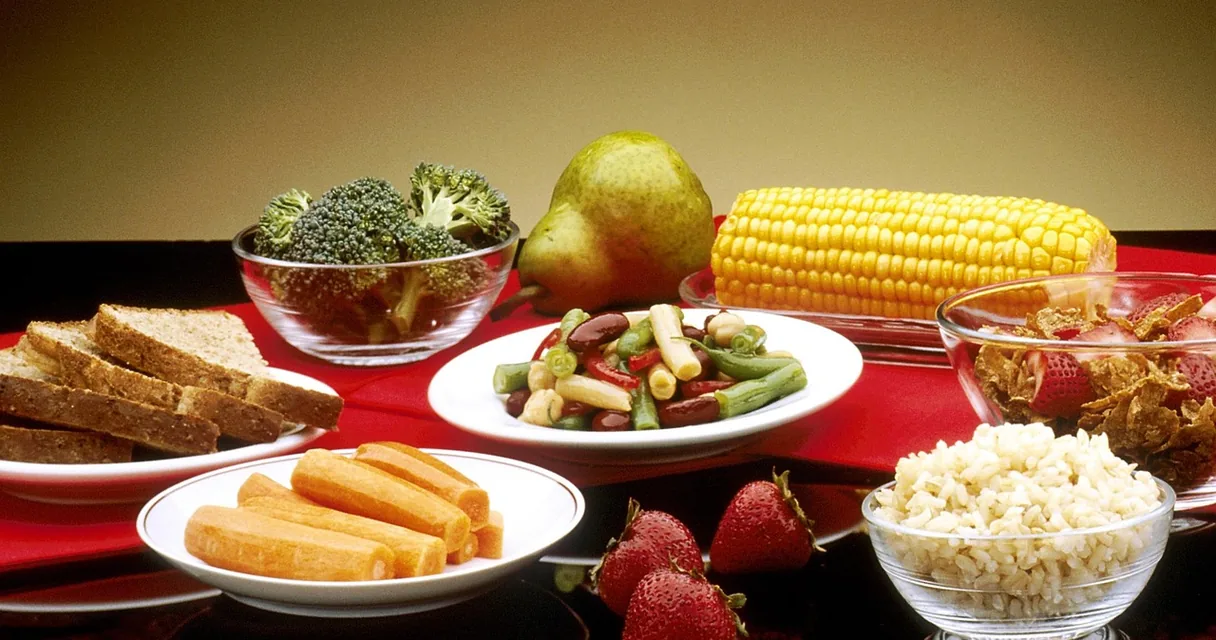 Mesa com pratos de legumes, milho, pães e frutas, ilustrando a diferença entre vegano e vegetariano