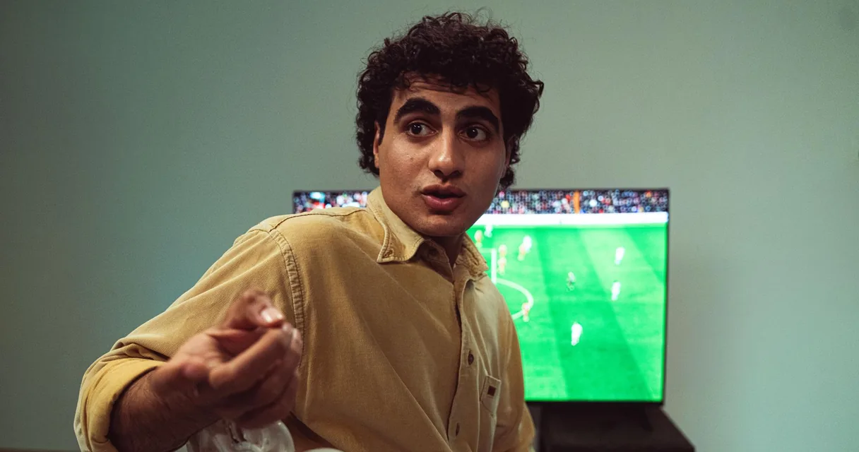 Homem de camisa mostarda diante de uma partida de futebol na televisão, ilustrando o futebol no celular